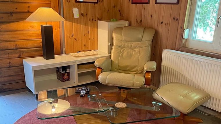Een gezellige woonruimte met een comfortabele stoel en een glastafel. De wanden zijn van hout en er is een warme gloed van een lamp.