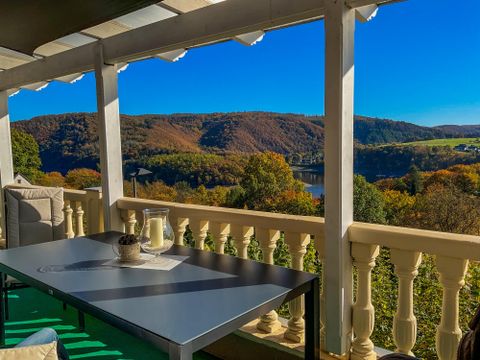 Eine gemütliche Terrasse mit einem Tisch und Sesseln bietet einen malerischen Blick auf die herbstliche Landschaft und den Fluss. Der klare blaue Himmel und die bunten Bäume sorgen für eine entspannte Atmosphäre.