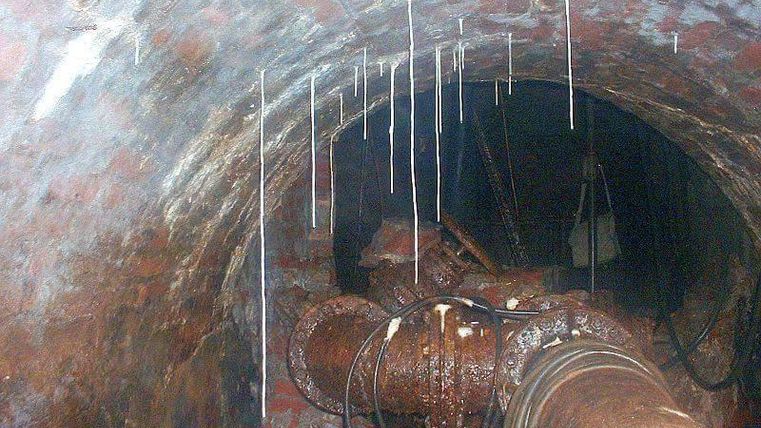 Vue d'un tunnel sombre aux murs en briques rouges. Des gouttelettes d'eau pendent aux murs et des tuyaux sont visibles.