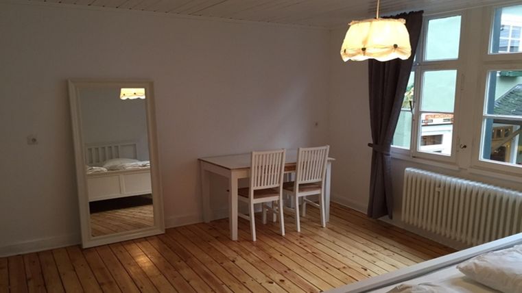 Une chambre simple et lumineuse avec une table et quatre chaises. Un grand miroir est accroché au mur et les fenêtres laissent entrer suffisamment de lumière naturelle.