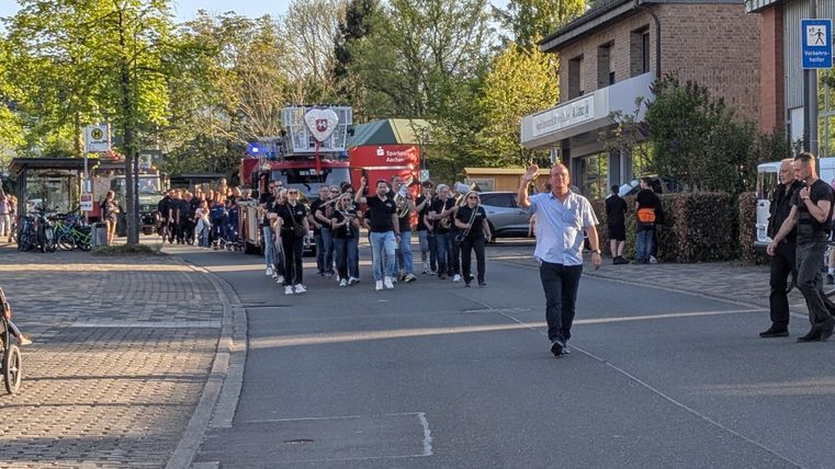 Eine Gruppe von Menschen marschiert auf einer Straße, während ein Mann in weißem Hemd ihnen entgegenkommt. Im Hintergrund sind Fahrzeuge und Gebäude sichtbar.