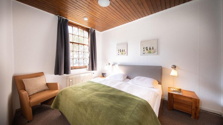 Ein gemütliches Schlafzimmer mit einem Doppelbett und einem Grün überzogenen Bettlaken. Es gibt einen Lesesessel und große Fenster, die für natürliches Licht sorgen.