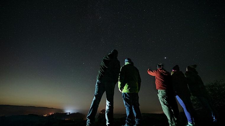 Eine Gruppe von Menschen steht im Dunkeln und schaut in den Nachthimmel. Sterne glänzen klar am Himmel.