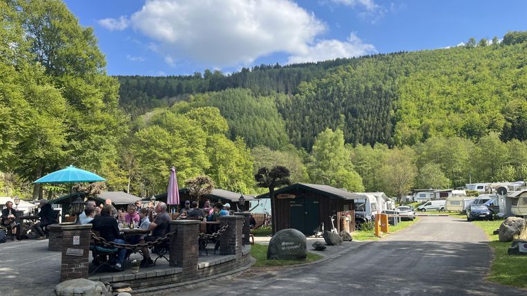 Ein Campingplatz in einer idyllischen Umgebung mit grünen Hügeln. Mehrere Wohnmobile sind auf dem Gelände, und eine Gruppe von Menschen sitzt entspannt im Freien.