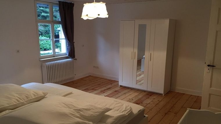 Une chambre simple avec un grand lit et un parquet en bois. Une armoire claire et une fenêtre avec des rideaux créent une atmosphère agréable.