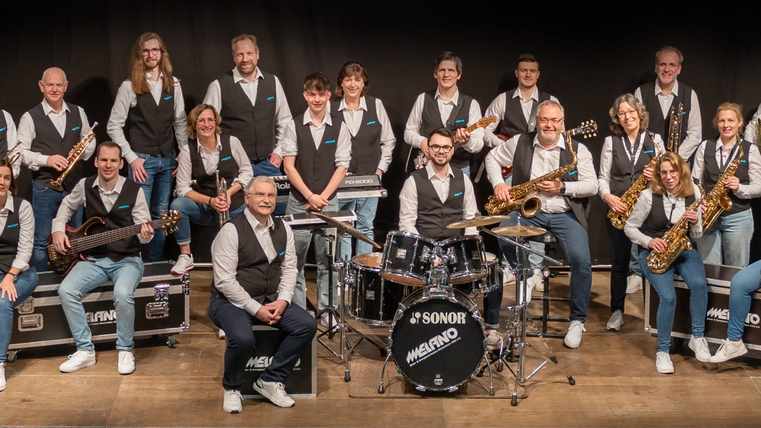 Eine große Band steht auf einer Bühne und posiert zusammen. Die Mitglieder tragen einheitliche Kleidung und halten verschiedene Musikinstrumente.