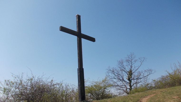 A black cross stands on a hill. In the background, there is a clear blue sky and some plants visible.
