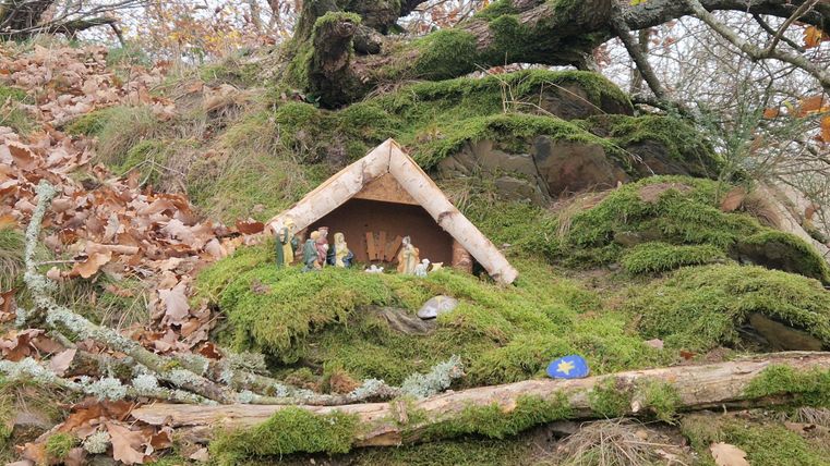 Eine kleine Hütte aus Holz steht auf moosbedecktem Boden, umgeben von buntem Herbstlaub. Vor der Hütte sind Figuren platziert, die eine Szene darstellen.