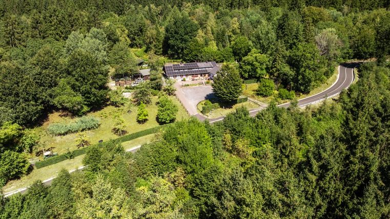 Eine ruhige Landschaft mit einem großen, umgebenen Wald und einer Straße, die durch die Natur führt. Im Vordergrund ist ein Gebäude mit einem begrünten Grundstück zu sehen.