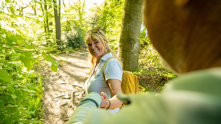 Een vrouw glimlacht op een zonnig bospad en houdt de hand van een ander vast. De omgeving is groen en vol bomen.