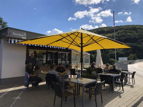 Ein gemütliches Café im Freien mit einem großen gelben Sonnenschirm. Gäste sitzen an Tischen und genießen die Sonne.