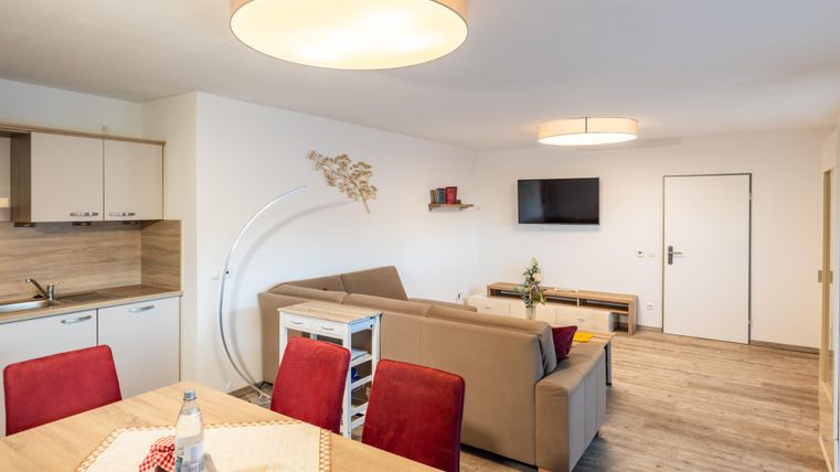 Ein modernes Wohnzimmer mit einer gemütlichen Couch und einem Esstisch. Die Wände sind hell, und es gibt einen Fernseher sowie einige Dekorationselemente.