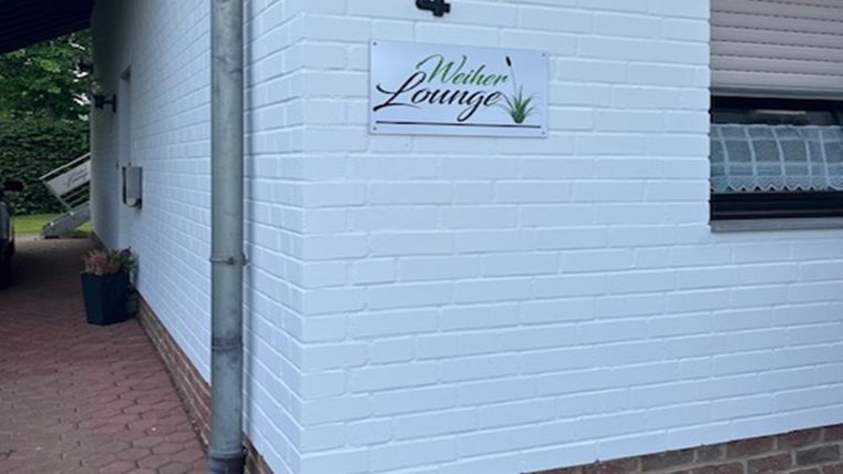 Ein weißes Haus mit einer hellen Wand und einer kleinen Schilder mit der Aufschrift „Weberei Lounge“. Neben dem Eingang steht ein grünes Pflanzgefäß.