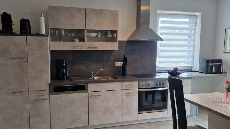 Eine moderne Küche mit grauen Schränken und Edelstahlgeräten. Der Raum ist hell und einladend, mit einem Esstisch und einer großen Fenstersonne.