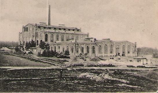 Ein historisches Fabrikgebäude mit großen Fenstern und einem Schornstein. Es befindet sich in einer ländlichen Umgebung mit einigen Baustellen und unbefestigten Flächen.