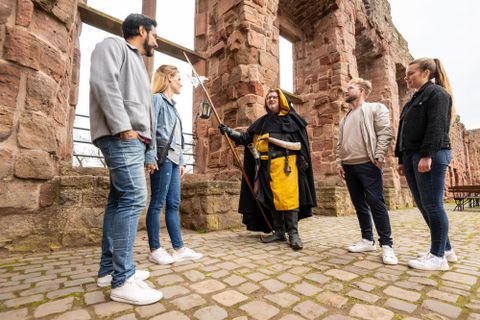 Eine Gruppe von fünf Personen steht in einer alten Ruine. Ein Mann in einem Kostüm erzählt etwas mit einem Zauberstab in der Hand.