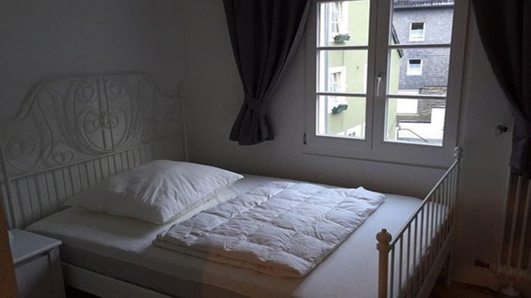 Une chambre simple avec un lit blanc et une fenêtre. Les murs sont clairs et il y a des rideaux qui atténuent doucement la lumière.