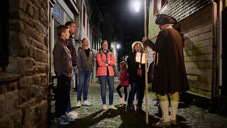 Eine Gruppe von Menschen hört einem Geschichtenerzähler in historischer Kleidung in einer schmalen, beleuchteten Gasse zu. Es ist Nacht und die Atmosphäre ist freundlich und einladend.