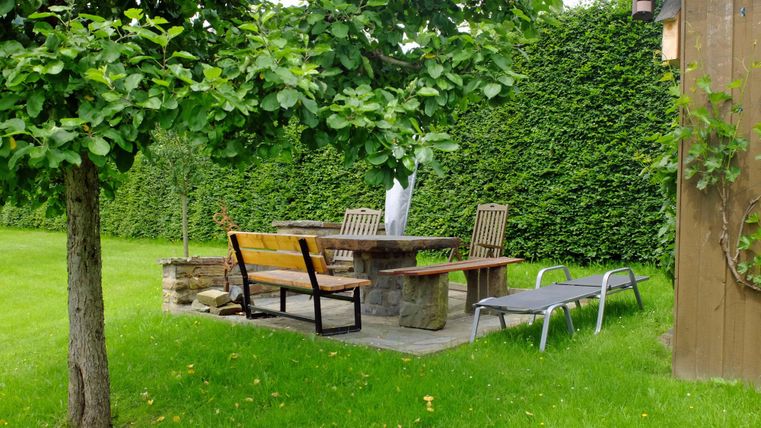 Ein einladender Garten mit einer Steintischgarnitur und mehreren Stühlen. Die Umgebung ist von einer hohen Hecke umgeben und es gibt eine Liege zum Entspannen.