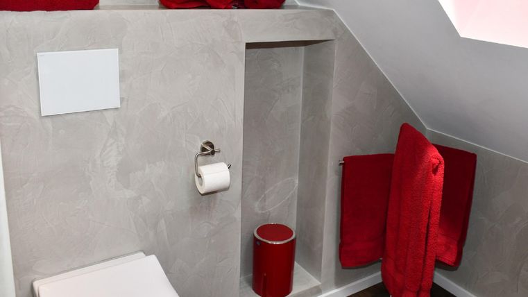 Une salle de bain moderne avec un plafond incliné et des murs gris. Des serviettes rouges et une tasse rouge apportent des touches de couleur.