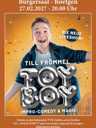 A cheerful man with a broad smile is promoting his new live show "Toy Boy". The event takes place on the 27th of February 2027 in the Bürgersaal Roetgen.