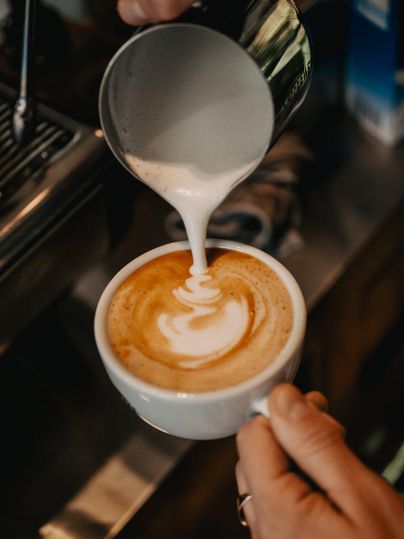 Eine Person schüttet dampfenden Milchschaum in eine Tasse Kaffee. Die kunstvolle Latte-Art ist deutlich sichtbar.