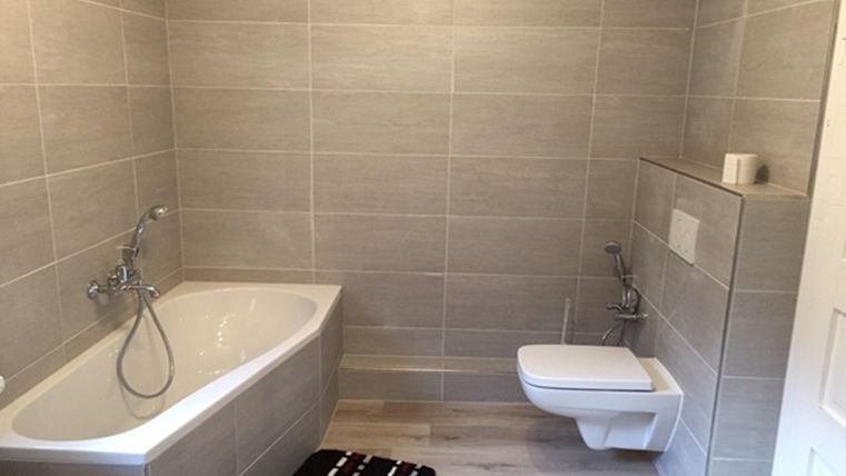 Une salle de bain moderne avec une baignoire et des toilettes. Les murs sont revêtus de carrelage gris et le sol est en bois.