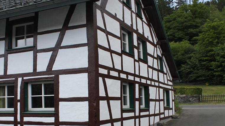 Ein traditionelles Fachwerkhaus mit weißen Wänden und dunklen Balken. Im Hintergrund sind Bäume und eine ruhige Umgebung zu sehen.