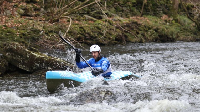 Ein Kajakfahrer paddelt energisch durch einen rauen Fluss. Um ihn herum fließt das Wasser und die Umgebung ist von Bäumen gesäumt.