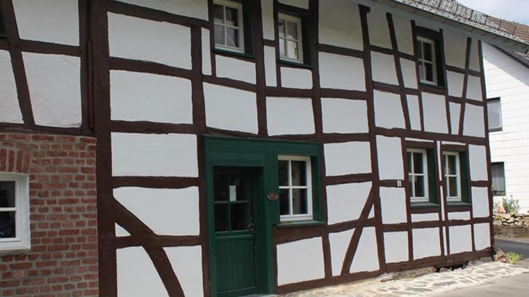 Ein traditionelles Fachwerkhaus mit weißen Wänden und braunen Balken. Die Tür ist grün und der Boden ist mit Steinen gepflastert.