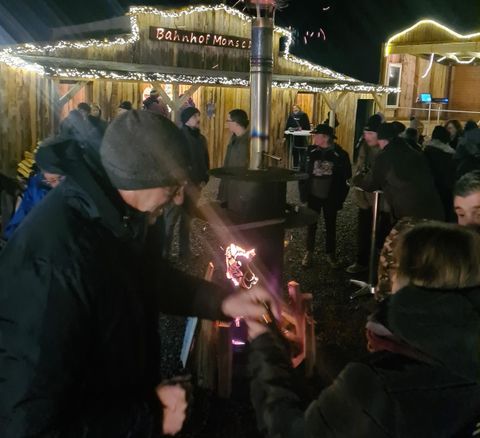 Eine festliche Szene mit Menschen, die rund um einen Holzstand stehen. Es ist dunkel, und die Umgebung ist mit Lichtern geschmückt.
