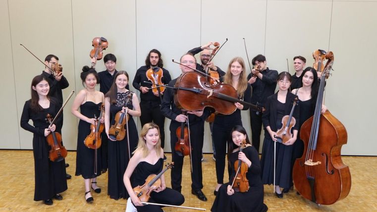 Ein Orchester mit Musikern in schwarzer Kleidung. Sie spielen verschiedene Streichinstrumente auf einer Bühne.