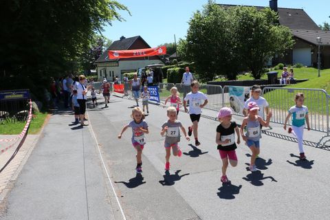 Eine Gruppe von Kindern läuft einen Wettlauf auf einer sonnigen Straße. Um sie herum stehen Zuschauer, und im Hintergrund ist eine Zielgerade mit einem Banner zu sehen.