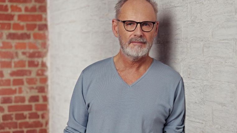 Ein Mann mit Brille steht vor einer hellen Ziegelwand. Er trägt einen hellblauen Pullover und hat graue Haare.