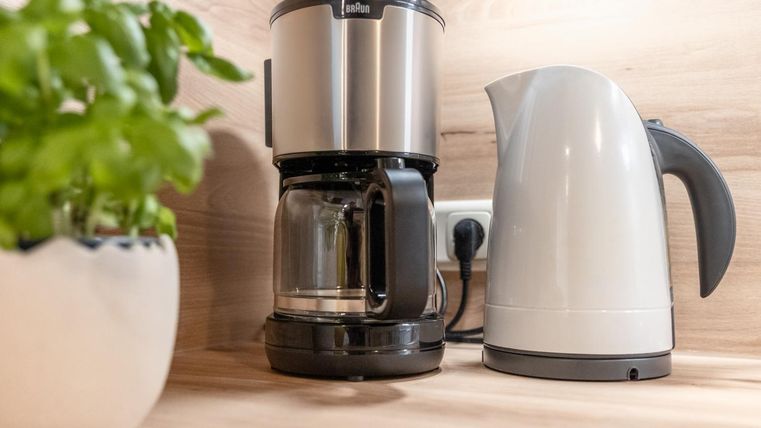Eine moderne Kaffeemaschine steht neben einem Wasserkocher auf einer Holzoberfläche. Im Hintergrund ist ein Topf mit frischen Kräutern zu sehen.