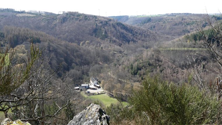 Eine malerische Aussicht auf ein weitläufiges Tal mit bewaldeten Hügeln. Im Tal sind einige Gebäude und Wiesen sichtbar.