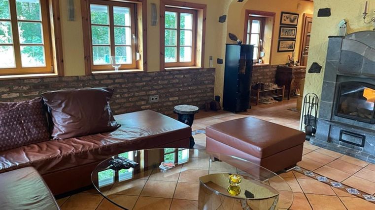 Een gezellige woonkamer met een L-vormige leren bank en een ronde glazen tafel. Grote ramen laten veel daglicht binnen en de open haard zorgt voor een warme sfeer.