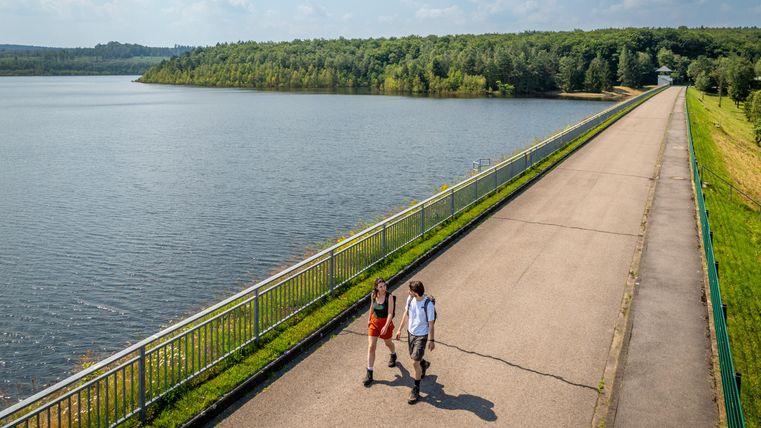 Twee personen wandelen langs een dijk naast een rustige meer. Op de achtergrond zijn bomen en een heldere, blauwe lucht te zien.