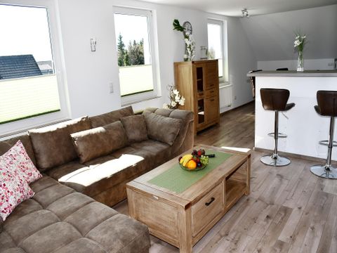Un salon lumineux avec un canapé beige et une table en bois. En arrière-plan se trouve une cuisine moderne.