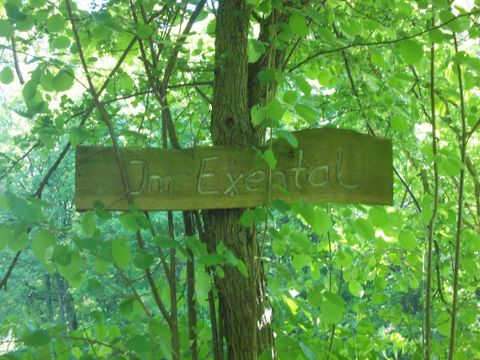 Ein Holzschild mit der Aufschrift "Im Exertal" hängt an einem Baum. Die Umgebung ist von grünen Blättern und Bäumen umgeben.