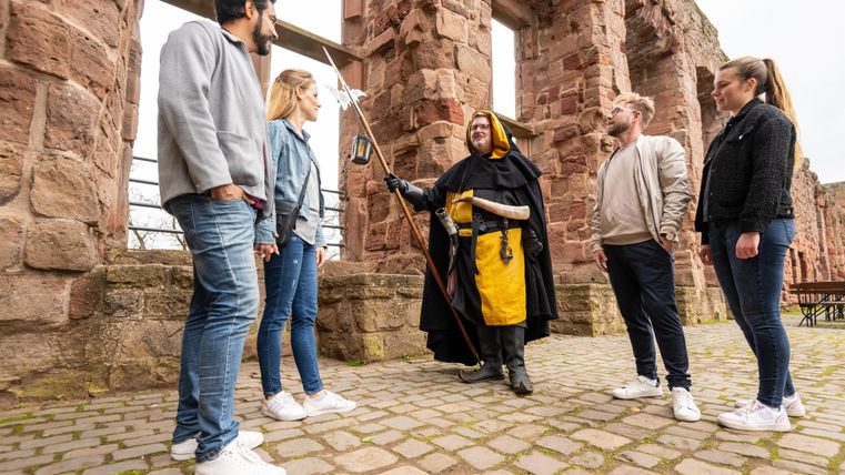 Eine Gruppe von fünf Personen steht in einer alten Ruine. Ein Mann in einem Kostüm erzählt etwas mit einem Zauberstab in der Hand.