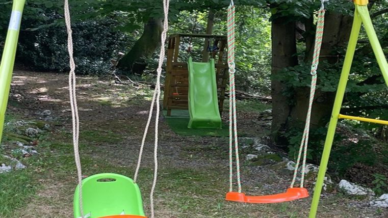 Een schommel met twee zitplaatsen staat in een groene, beboste omgeving. Op de achtergrond is een glijbaan te zien.