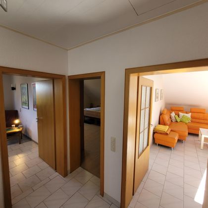 Ein weiträumiger Flur mit drei Türen, die zu verschiedenen Räumen führen. Im Hintergrund ist ein gemütliches Wohnzimmer mit einer orangefarbenen Couch sichtbar.