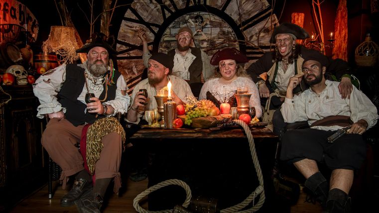 Een groep piraten zit om een tafel met voedsel en dranken. De sfeer is feestelijk en de omgeving is in piratenstijl gedecoreerd.