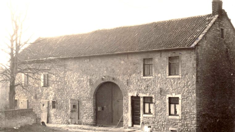 An old stone house with a large gate and several windows. In the foreground, you can see a few trees and a clear road.