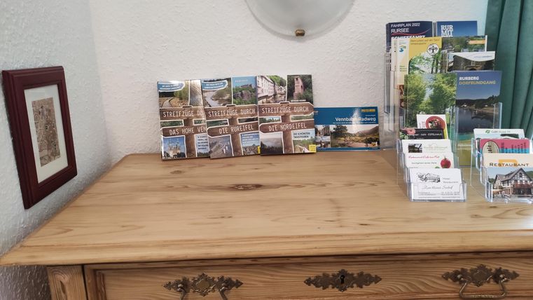 Ein holzfarbener Tisch mit einer kleinen Auswahl an Postkarten und Informationsmaterialien. Im Hintergrund hängt ein gerahmtes Bild an der Wand.