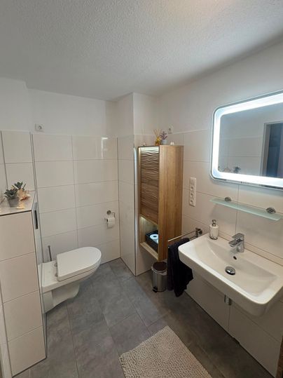 Une salle de bain moderne avec des carreaux clairs et une douche spacieuse. Il y a un lavabo, des toilettes et une armoire en bois pour un espace de rangement supplémentaire.