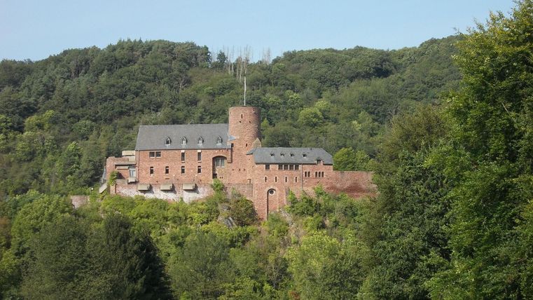 An old castle, surrounded by green forests and hills. The sky is clear and blue.