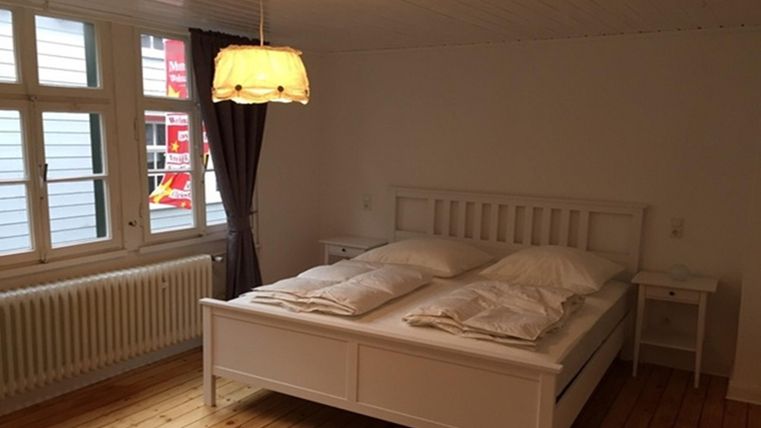 Une chambre lumineuse avec un lit blanc et deux coussins. De grandes fenêtres et un éclairage décoratif créent une atmosphère accueillante.
