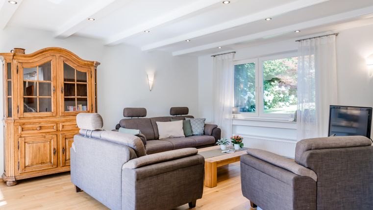 Ein gemütliches Wohnzimmer mit einem grauen Sofa und zwei Sesseln. Zu sehen sind auch ein Holzschrank und ein Fenster mit Blick ins Grüne.